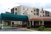Exterior view of Fellowship at The Fairway Assisted Living Community showing a multi-story beige building with balconies, a green canopy over the entrance driveway, an American flag on a flagpole, and landscaped bushes in front.