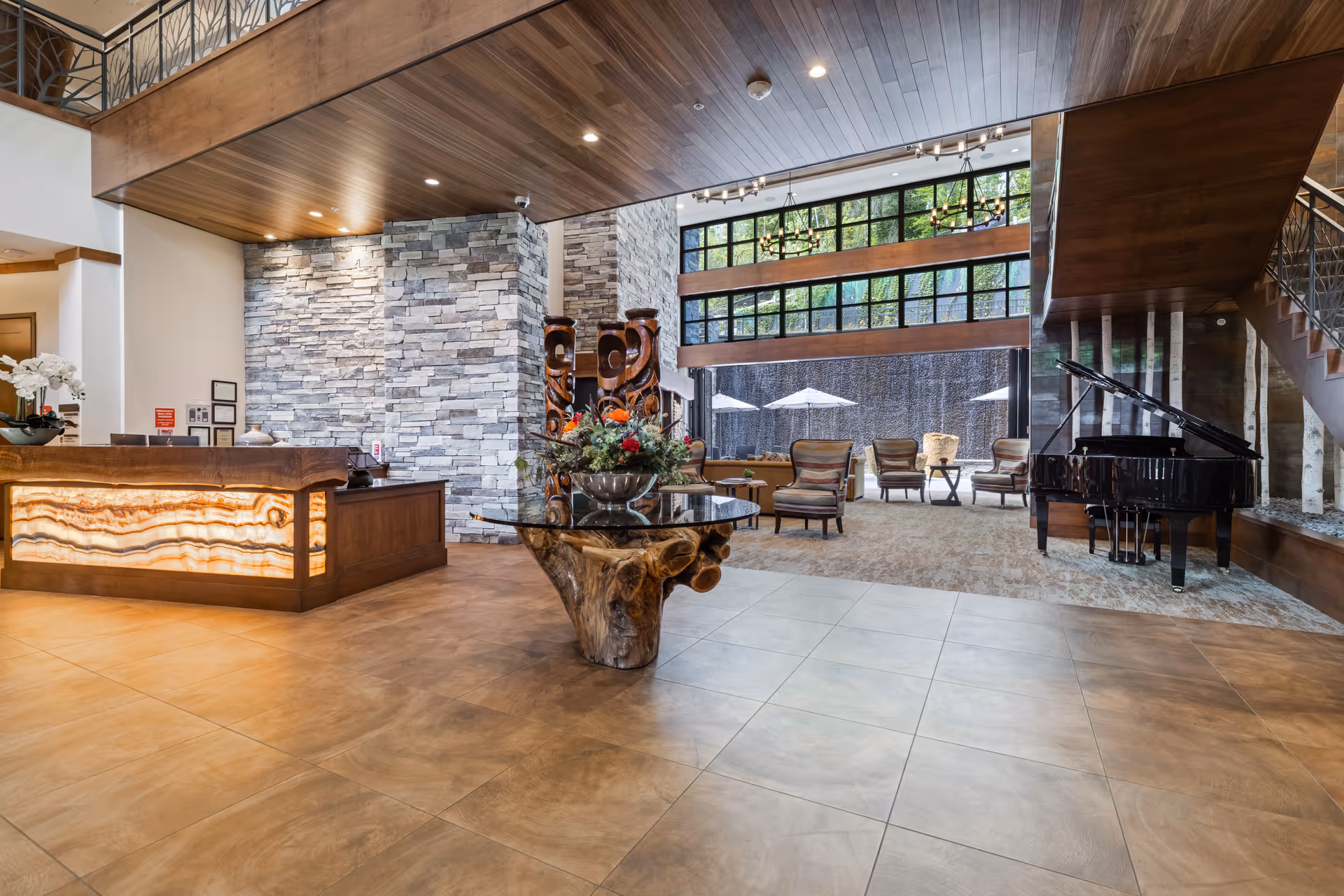 Spacious and elegant senior living facility lobby with a wooden reception desk featuring illuminated stone paneling, a large decorative wooden table with a floral arrangement, comfortable seating area with armchairs, a grand piano, stone accent walls, and large windows overlooking an outdoor patio with umbrellas.