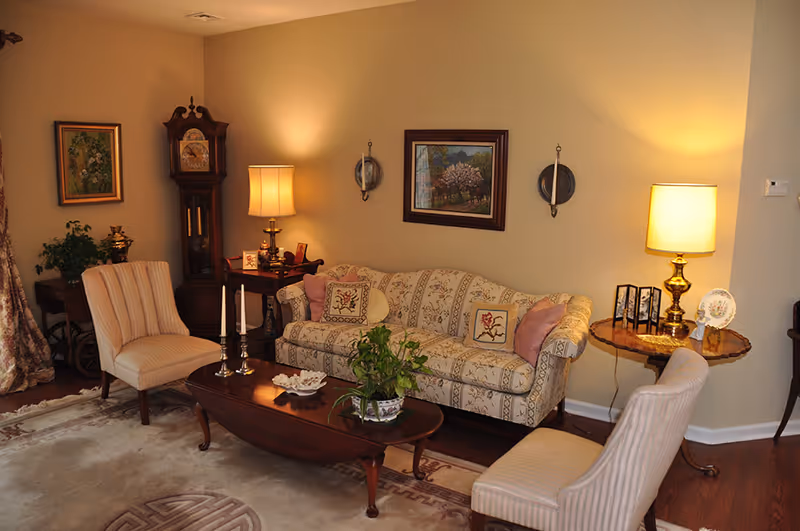 A cozy living room with a patterned sofa adorned with decorative pillows, two striped armchairs, a wooden coffee table with plants and candles, a grandfather clock, two table lamps, framed paintings on the walls, and a wooden side table with decorative items.