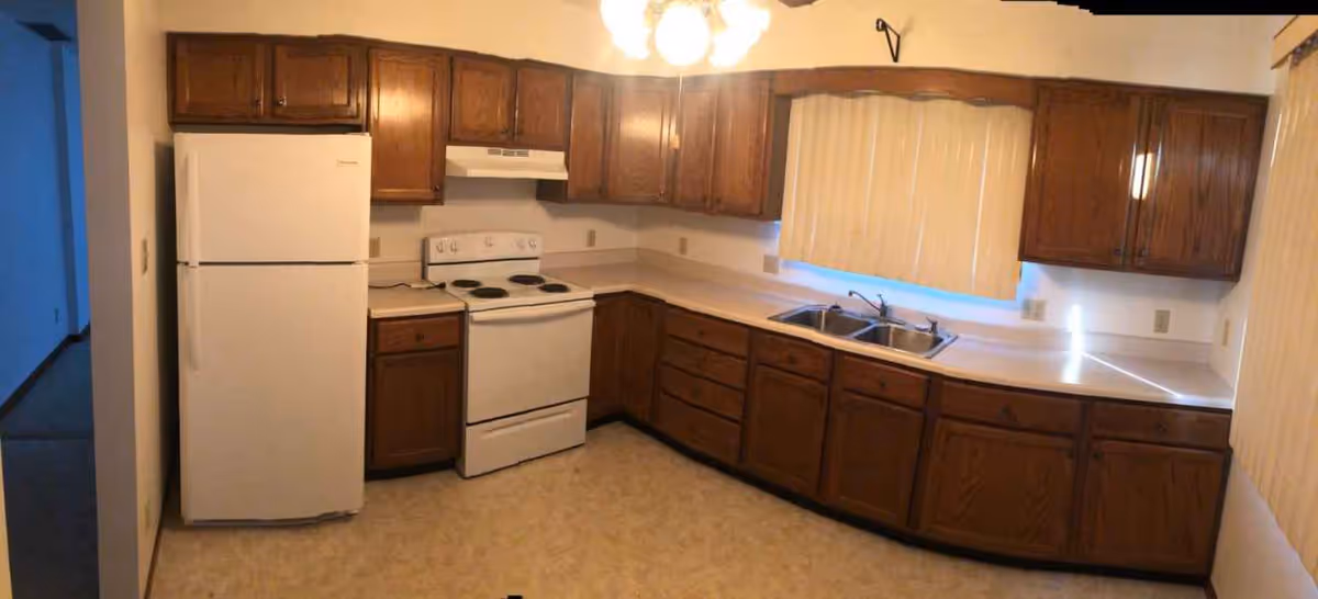 A kitchen with wooden cabinets, a white refrigerator, a white electric stove with an overhead vent, a double sink under a window with closed blinds, and beige countertops. The floor has a light-colored pattern, and there is a ceiling light fixture above.