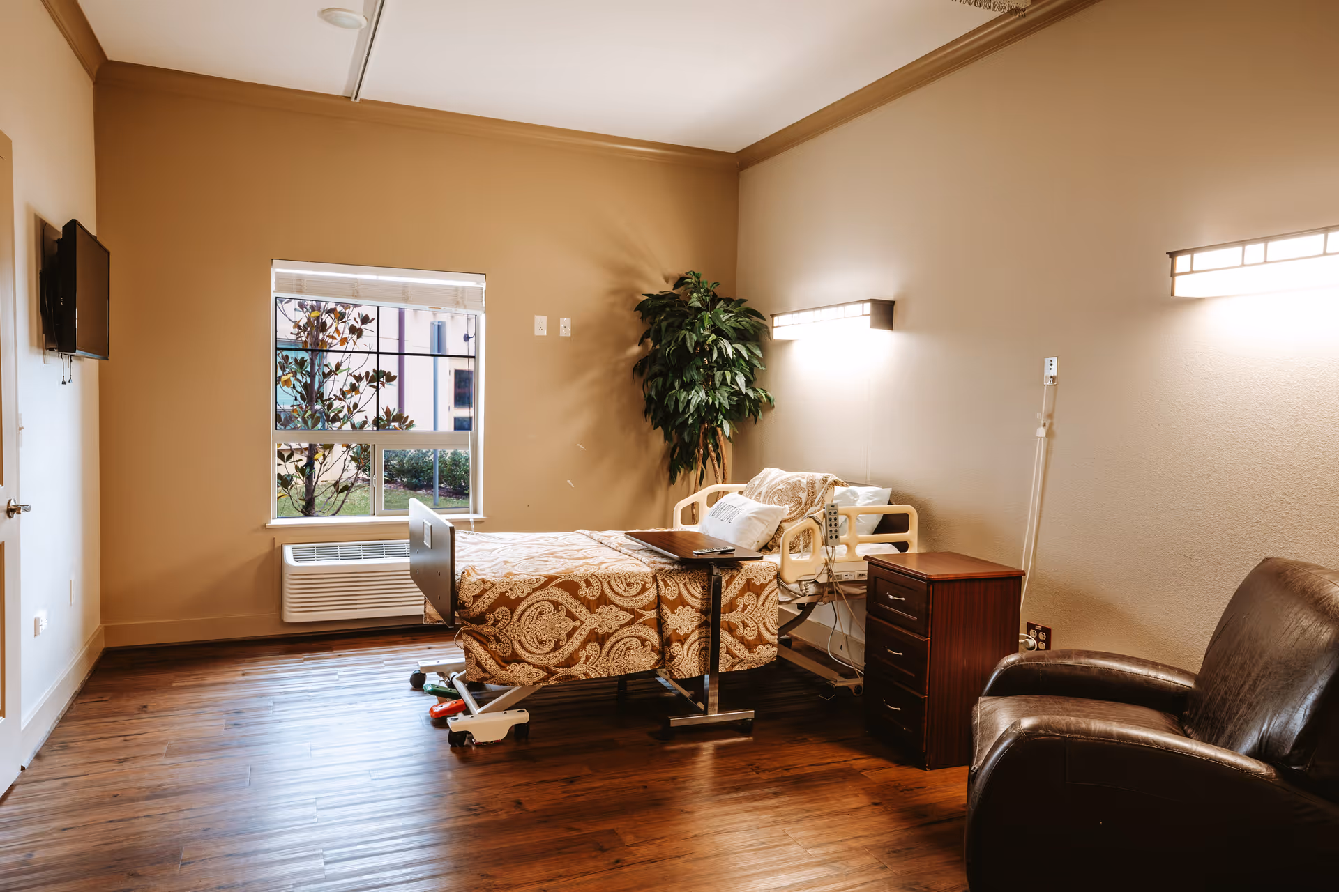 A nursing facility room with a hospital bed covered in a patterned blanket, a wooden bedside table with drawers, a brown leather armchair, a potted plant in the corner, a wall-mounted TV, and a window showing greenery outside. The room has wooden flooring and beige walls with two wall-mounted lights.