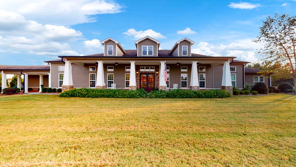 Front exterior view of Orchard Ridge Residences, a single-story building with a wide porch supported by white columns, three dormer windows on the roof, an American flag near the entrance, and a well-maintained lawn in the foreground under a partly cloudy sky.