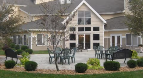 Outdoor patio area with several green plastic chairs and tables, surrounded by small bushes and trees, in front of a beige building with large windows and a peaked roof.
