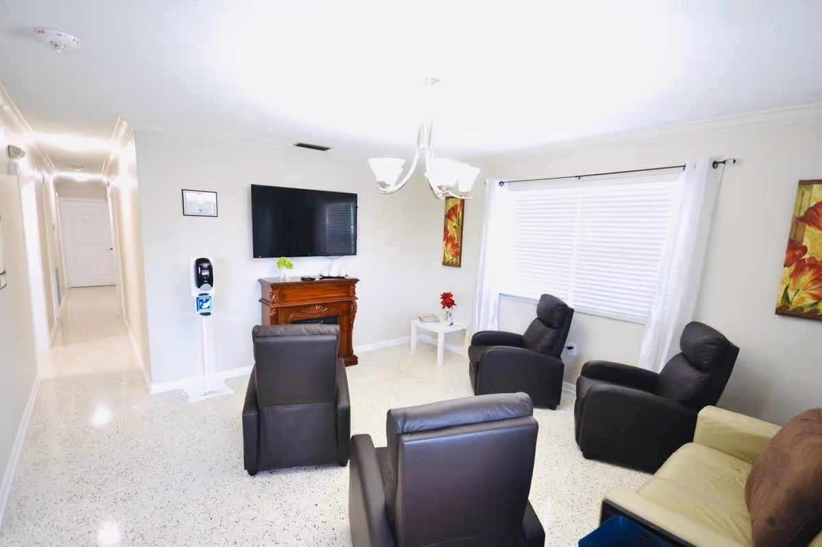 A bright living room with four black recliner chairs arranged around a wall-mounted flat screen TV above a wooden mantel. There is a small white side table with a red flower vase near a large window with white blinds and curtains. The floor is speckled, and the walls are painted white. A hallway is visible to the left.