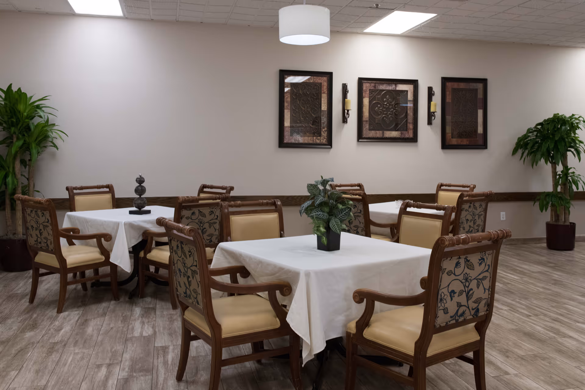 Dining room with several tables draped in white cloths, wooden chairs, potted plants, and framed artwork on the wall.
