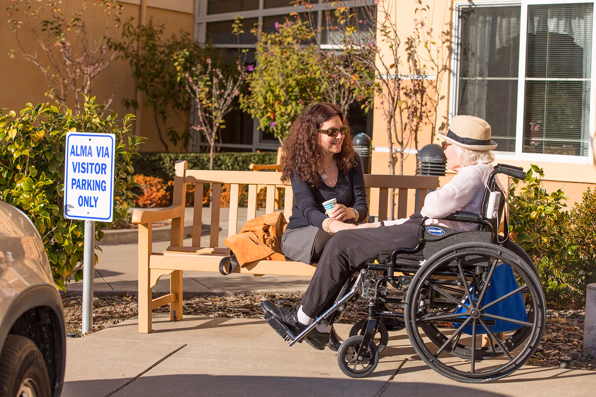 An elderly person in a wheelchair wearing a hat is sitting outside near a wooden bench, talking with a woman with curly hair and sunglasses who is sitting on the bench. There is a sign nearby that reads 'ALMA VIA VISITOR PARKING ONLY' and some greenery and a building with windows in the background.