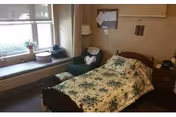 A cozy bedroom in a senior living facility with a single bed covered in a floral bedspread, a green armchair with a pillow, a wooden nightstand with a lamp and some items, a bulletin board on the wall, and a window seat with cushions and decorative items.