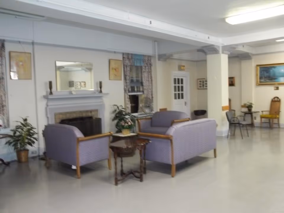 Spacious senior facility common room with two purple upholstered sofas arranged around a small table in front of a fireplace and additional chairs along the walls.