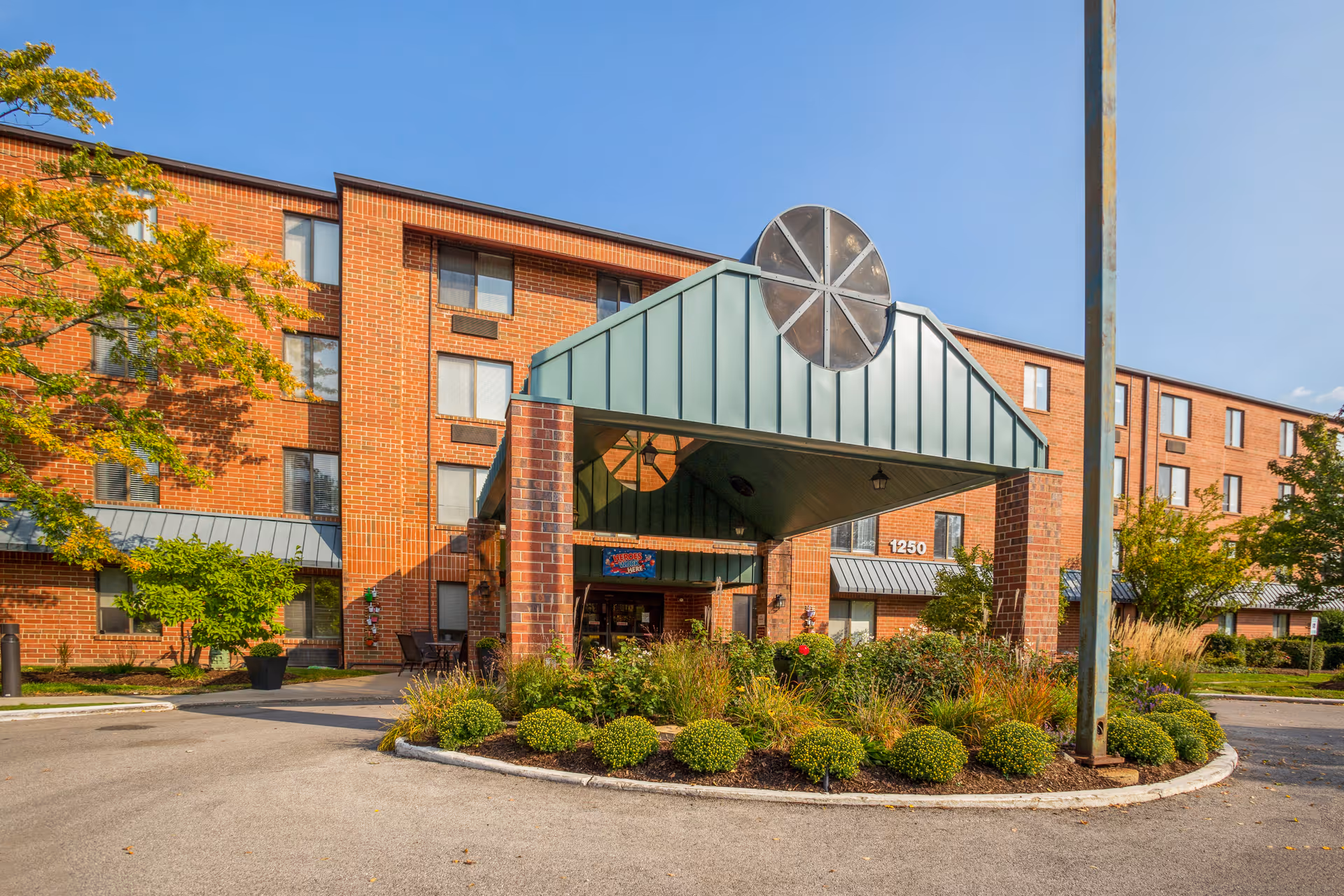Front entrance of a brick senior living building with a green covered porte-cochère, landscaped roundabout, and the address number 1250 visible.