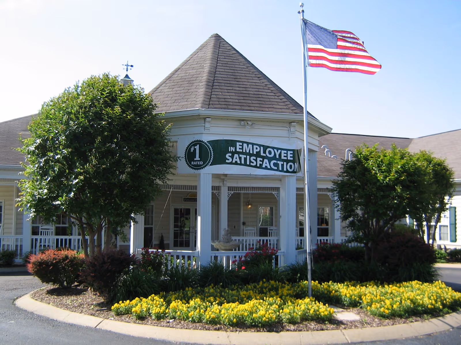 Exterior view of Charter Senior Living of Gallatin featuring a round gazebo-like entrance with a sign that reads '#1 Rated in Employee Satisfaction'. The building is surrounded by well-maintained landscaping including bushes, trees, and yellow flowers. An American flag is flying on a flagpole in front of the entrance.