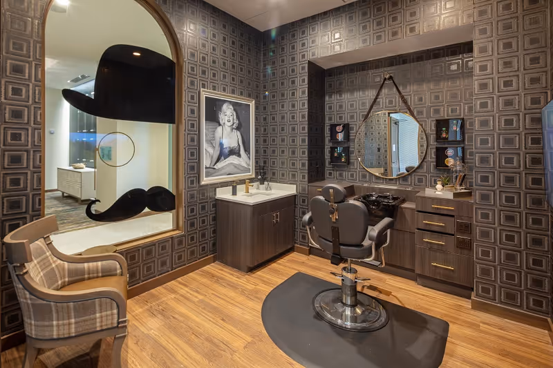 Interior view of a stylish salon room with patterned wallpaper, a barber chair in front of a vanity with a round mirror, a sink, and a cabinet. A framed black and white photo of Marilyn Monroe is on the wall. There is a plaid upholstered chair near a window with a decorative mustache and hat decal.