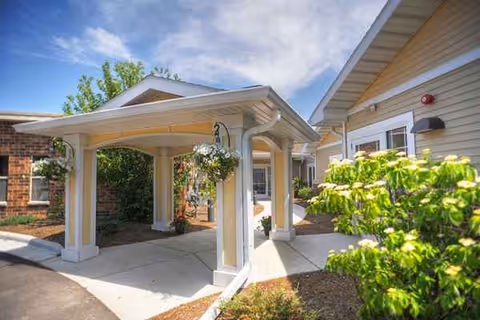 Covered entrance with columns leads to the building, surrounded by greenery and blue sky.