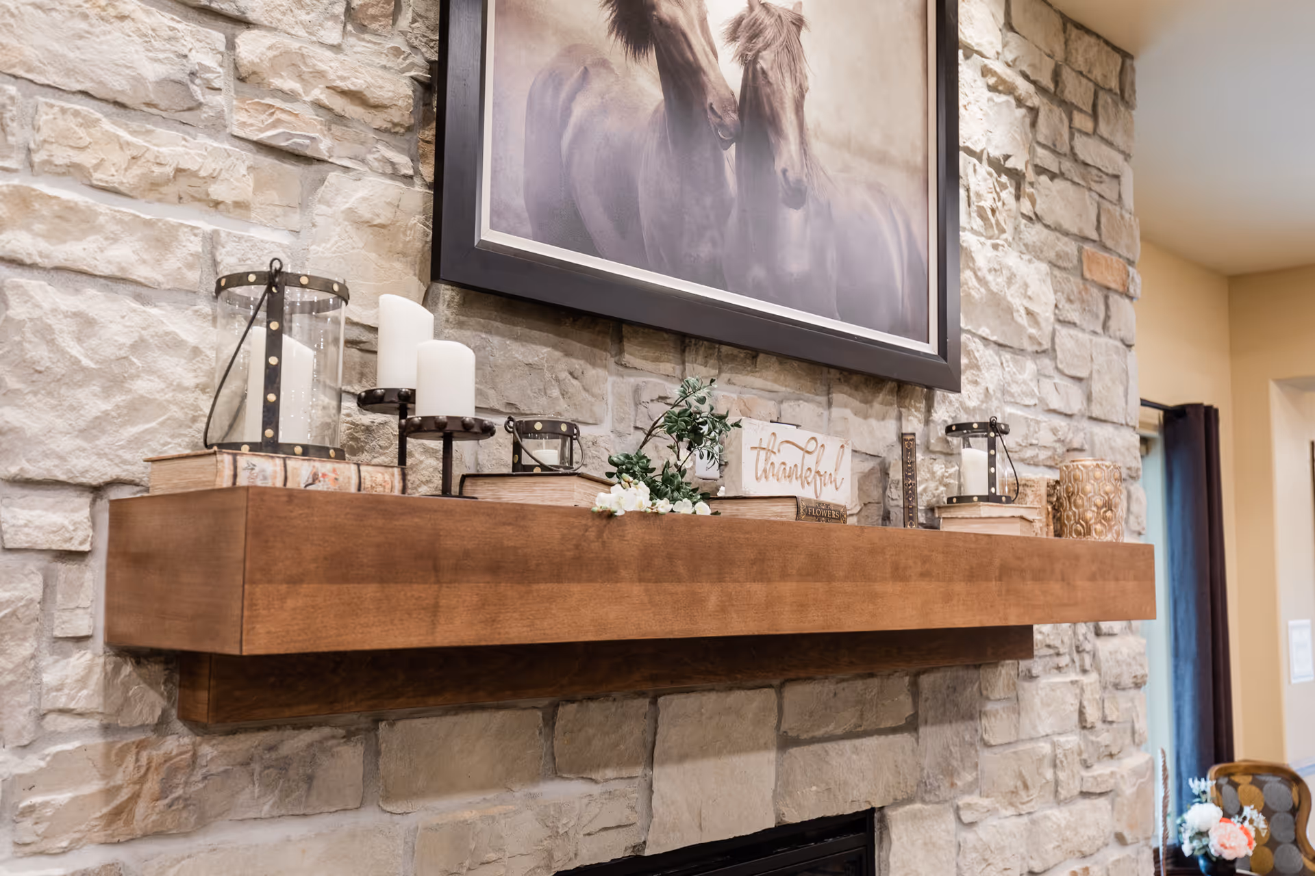A stone fireplace with a wooden mantel decorated with candles, books, a small plant, and a sign that says 'thankful'. Above the mantel is a framed picture of two horses. The room has a cozy and warm ambiance.