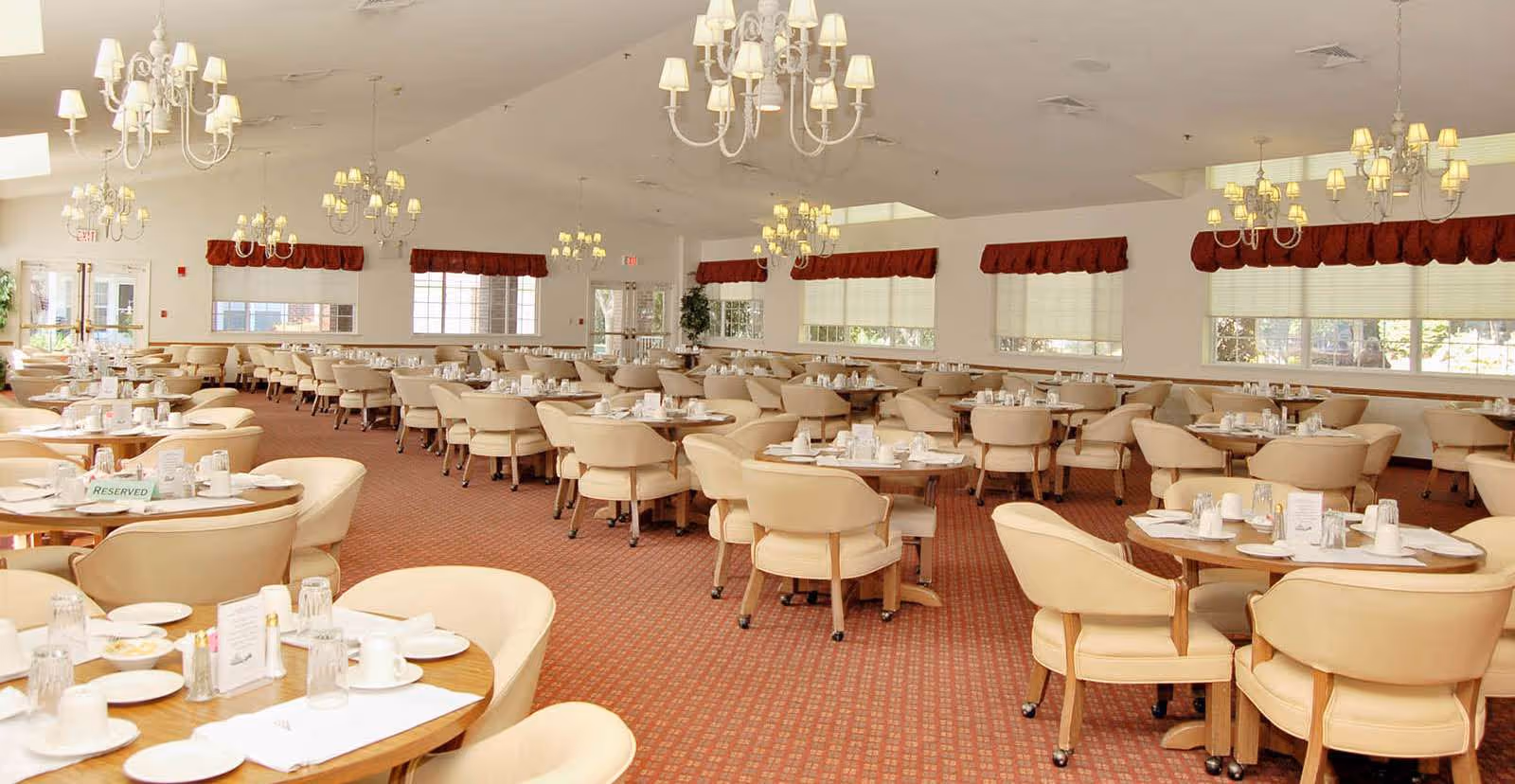 A large dining room with multiple round tables set with white plates, cups, and silverware. The room features beige cushioned chairs, red carpet flooring, white walls, large windows with red valances, and several chandeliers hanging from the ceiling providing warm lighting.