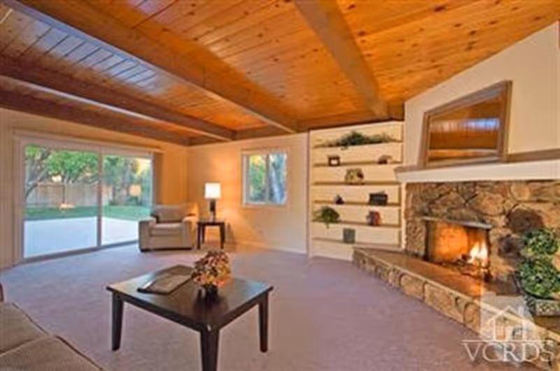 A cozy living room with a wooden beamed ceiling, a stone fireplace with a fire burning, built-in shelves with decorative items, a coffee table with a flower arrangement, an armchair with a side table and lamp, and large sliding glass doors leading to a green outdoor area.