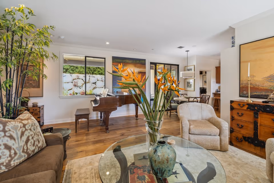 Bright, open living room with a grand piano, upholstered seating, a glass coffee table topped with flowers and decorative accents, and hardwood floors.