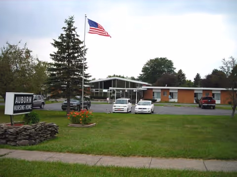 Exterior view of Auburn Nursing Home building with a lawn in front, an American flag on a flagpole, several parked cars, and a sign that reads 'Auburn Nursing Home'.