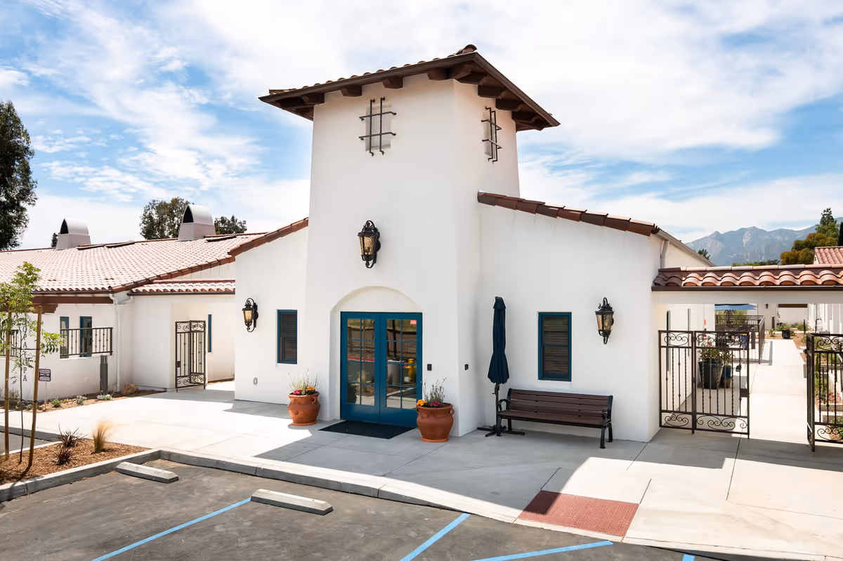 Front exterior of a white Spanish-style building with a red tile roof, central blue double doors, a bench, and potted plants.