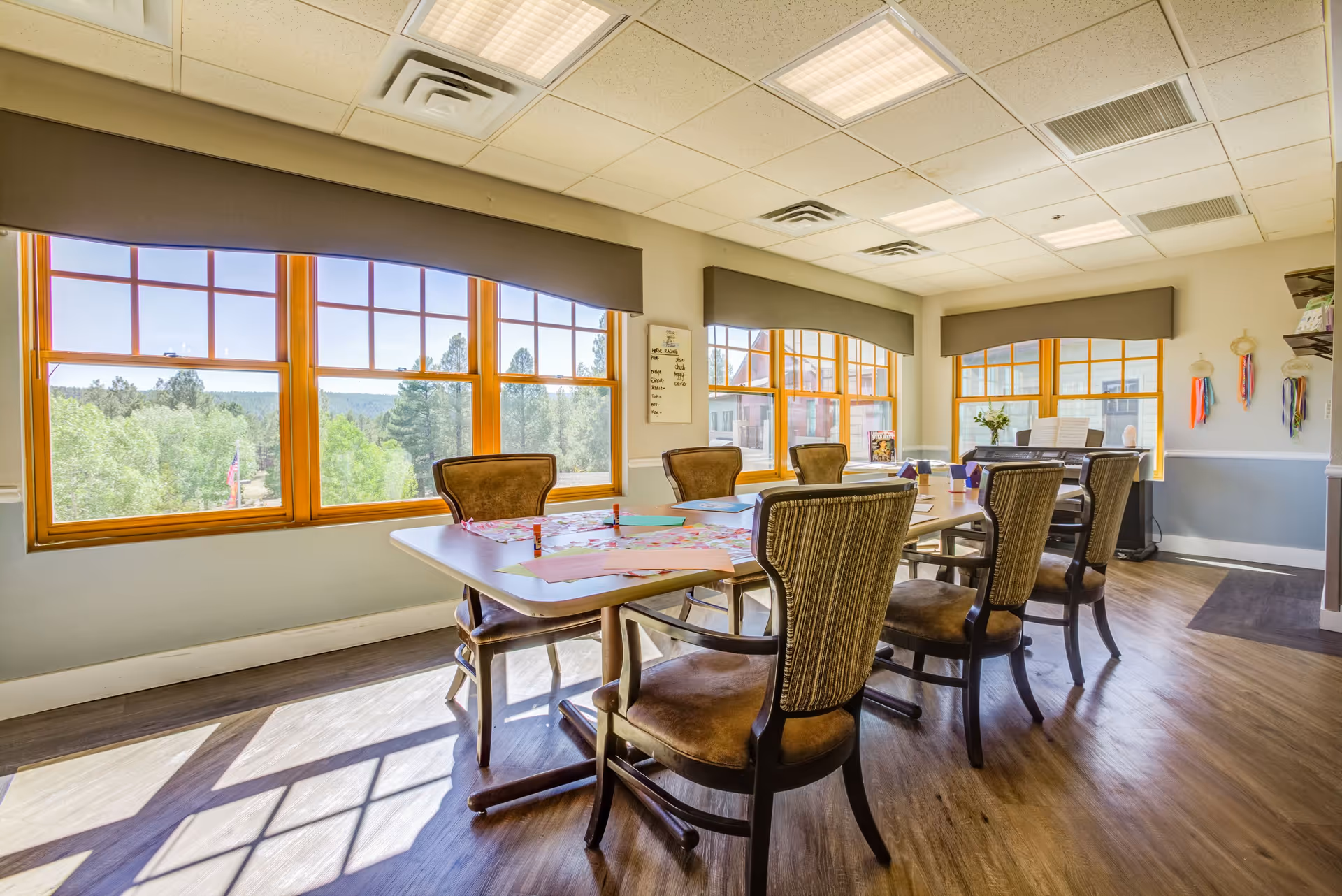 A bright and spacious room with large windows letting in natural light, featuring a long table surrounded by eight chairs. The table has various papers and craft supplies on it. The room has wooden flooring, a piano in the corner, and walls decorated with colorful ribbons and a small whiteboard.