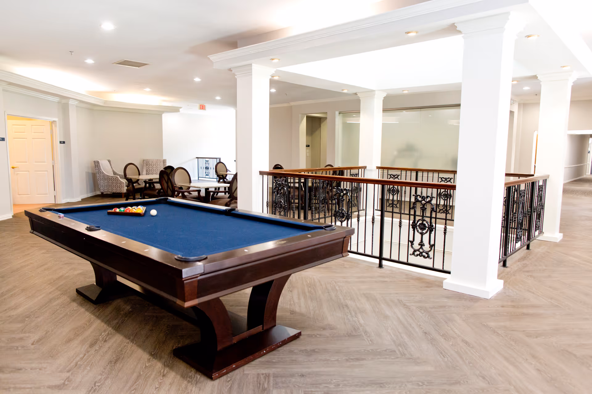 A spacious indoor common area featuring a pool table with blue felt in the foreground. Behind the pool table, there are several chairs and small tables arranged for seating. The space is well-lit with recessed lighting and has white walls and columns, with decorative black wrought iron railings around an open area. The flooring is a light wood pattern.