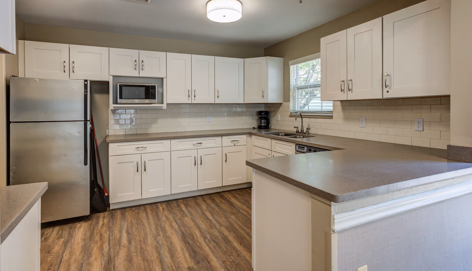 A modern kitchen with white cabinets, a stainless steel refrigerator, a built-in microwave, a coffee maker, a dishwasher, and a double sink under a window. The kitchen has a light brown countertop and wood-patterned flooring.