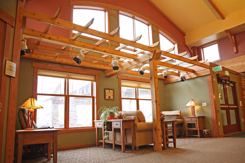 A cozy interior common area with wooden beams and large windows letting in natural light. The room features comfortable armchairs, side tables with lamps, and a potted plant. The walls are painted green and the ceiling is vaulted with exposed wooden beams.