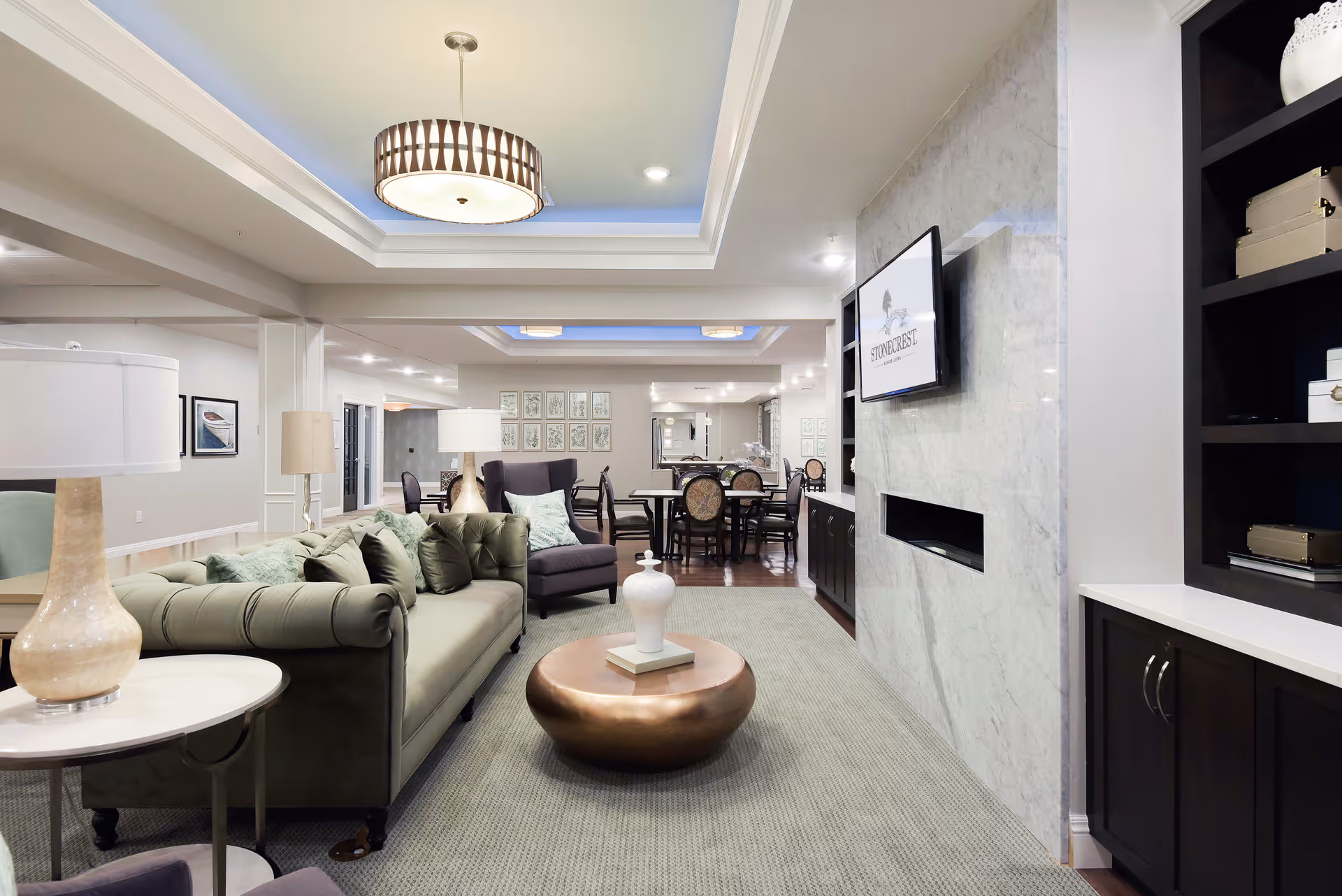 A modern and spacious living room area in a senior living facility featuring a green tufted sofa with multiple cushions, a round copper coffee table with a decorative vase, a dark armchair, and a flat-screen TV mounted on a marble wall above a built-in fireplace. The room has recessed lighting, a ceiling light fixture, and a dining area with tables and chairs visible in the background.