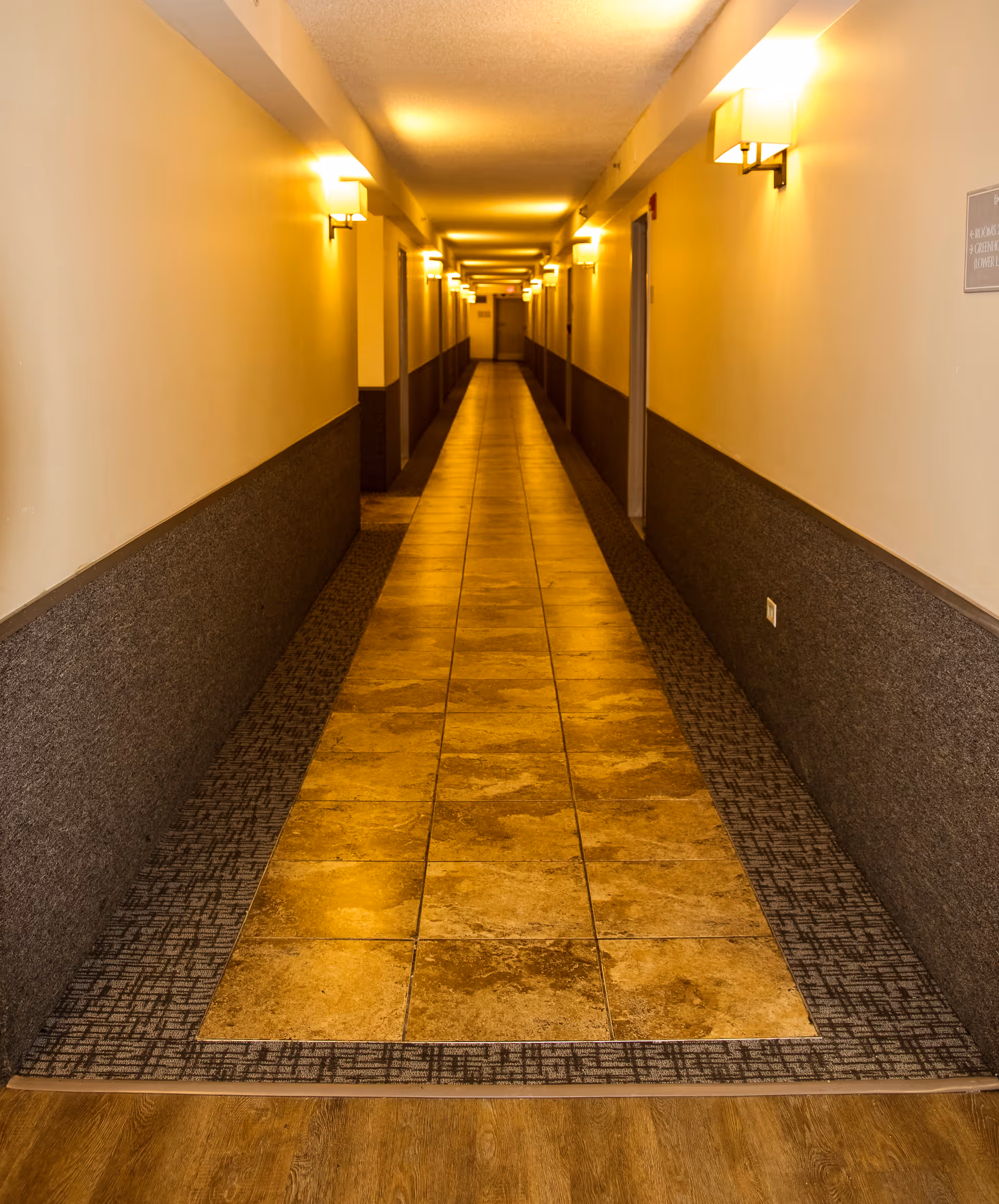 A long, narrow hallway in a building with tiled flooring in the center and carpeted edges. The walls are painted white on the upper half and dark gray on the lower half. Wall-mounted lights evenly illuminate the corridor, and several doors line both sides of the hallway.