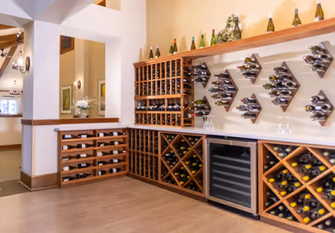 Interior view of a wine storage area with multiple wooden wine racks filled with bottles. There is a wine cooler in the center and decorative wine bottles displayed on a shelf above. The room has light-colored walls and wooden flooring.