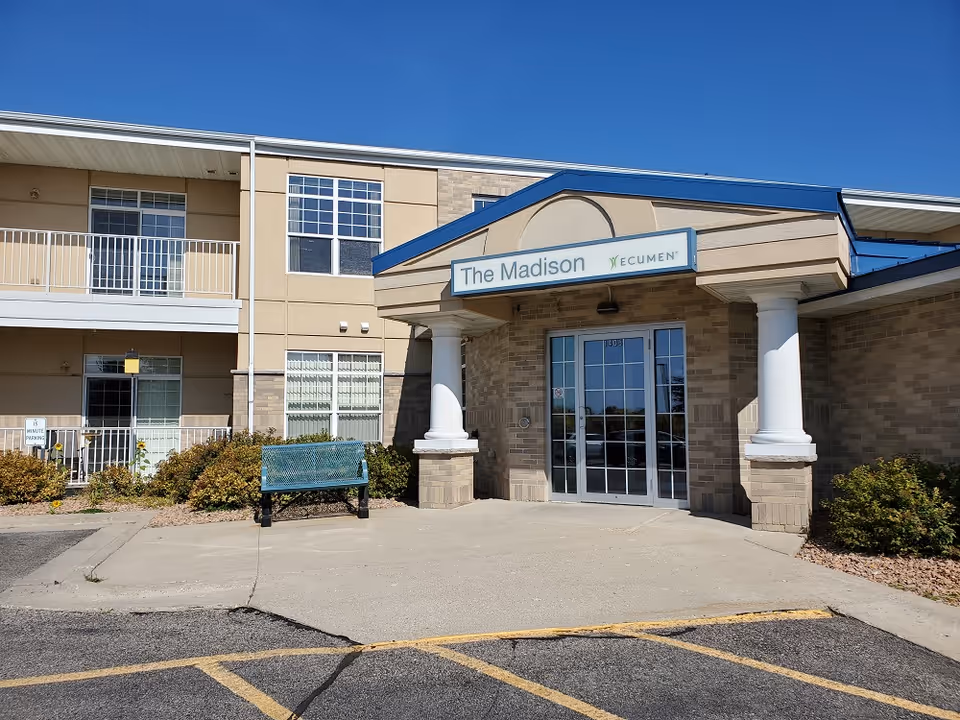 Front entrance of 'The Madison' senior living building with columns, glass double doors, and a bench outside.