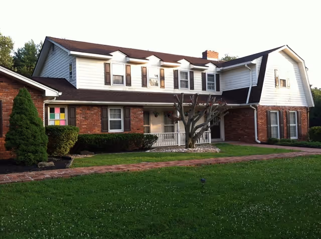 A two-story residential building with a combination of red brick and white siding exterior. The house features multiple windows with brown shutters, a small front porch with white railing, a trimmed tree in front, and a well-maintained green lawn with a brick pathway leading to the entrance.
