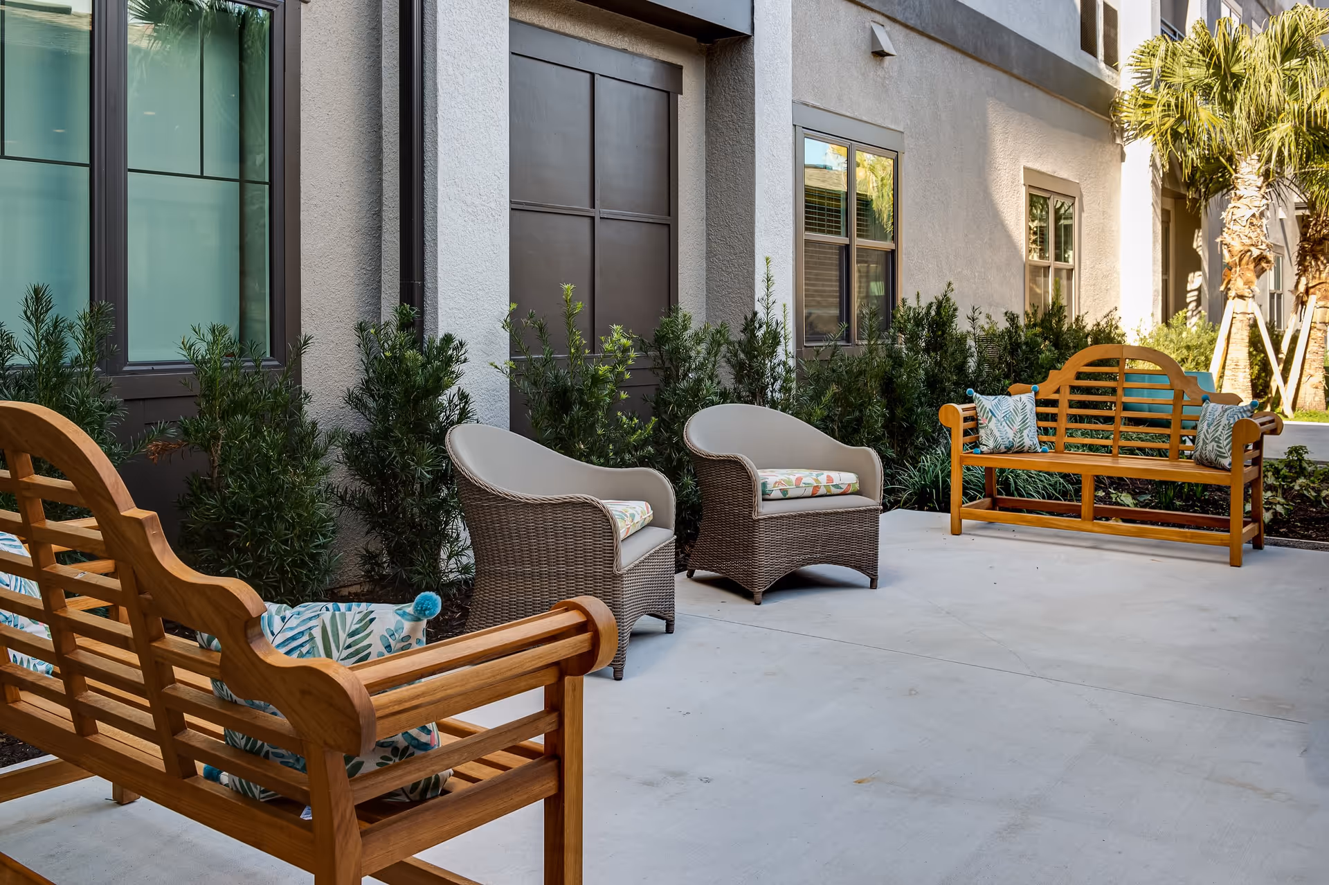 Outdoor patio area with two wooden benches and two wicker armchairs with cushions, surrounded by green shrubs and palm trees next to a building with windows.