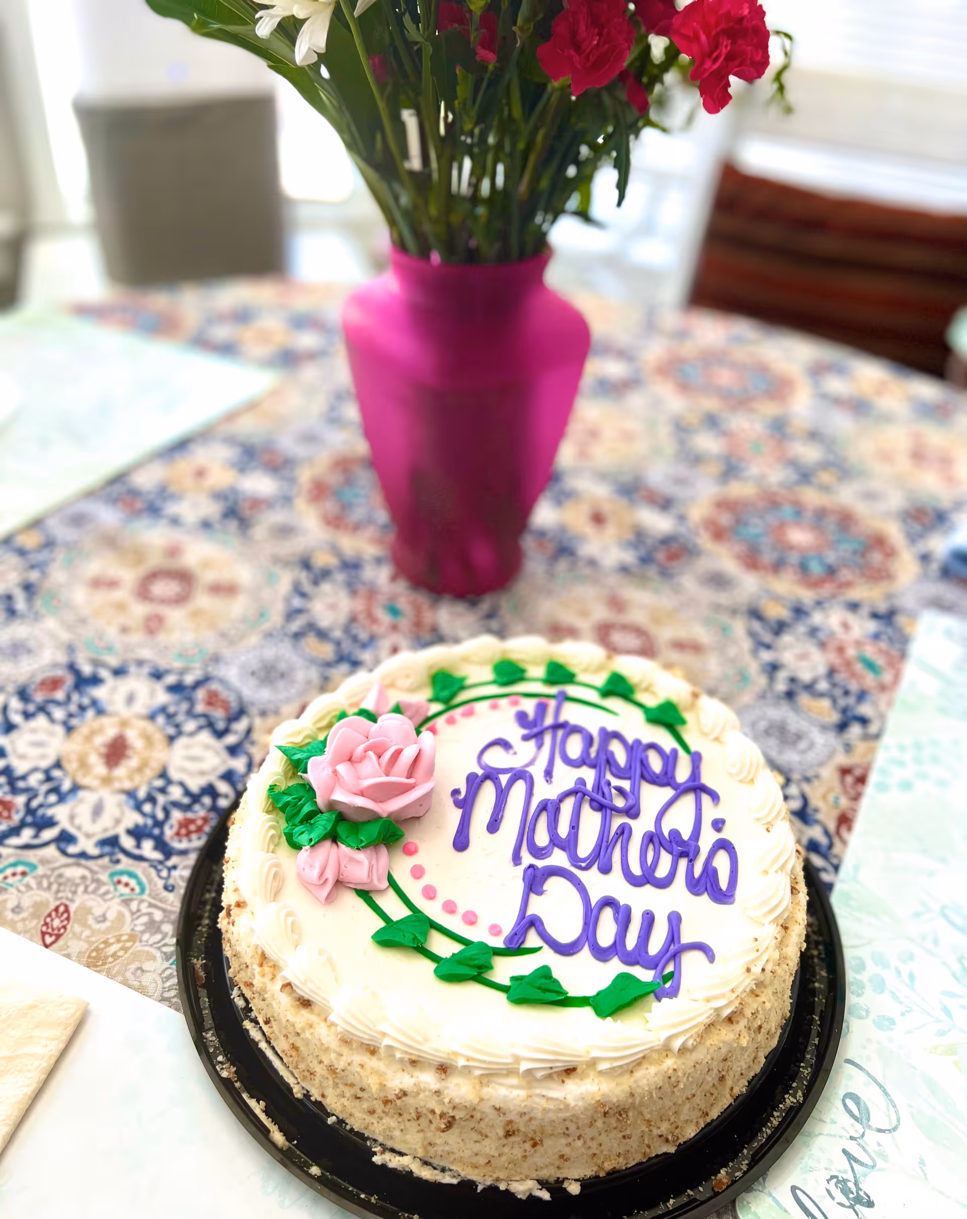 A round cake with white frosting decorated with a pink rose and green leaves, with the words 'Happy Mother's Day' written in purple icing, placed on a patterned tablecloth. Behind the cake is a pink vase with white and red flowers.