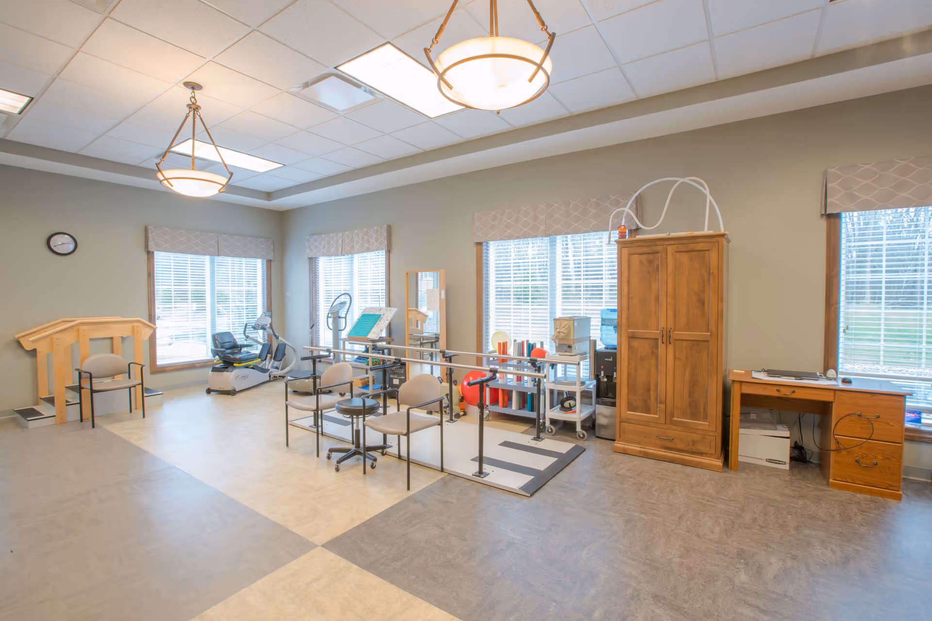 A bright physical therapy or rehabilitation room with exercise equipment including a stationary bike, parallel bars for walking practice, chairs, a wooden cabinet, and a desk. Large windows with blinds allow natural light to fill the space.