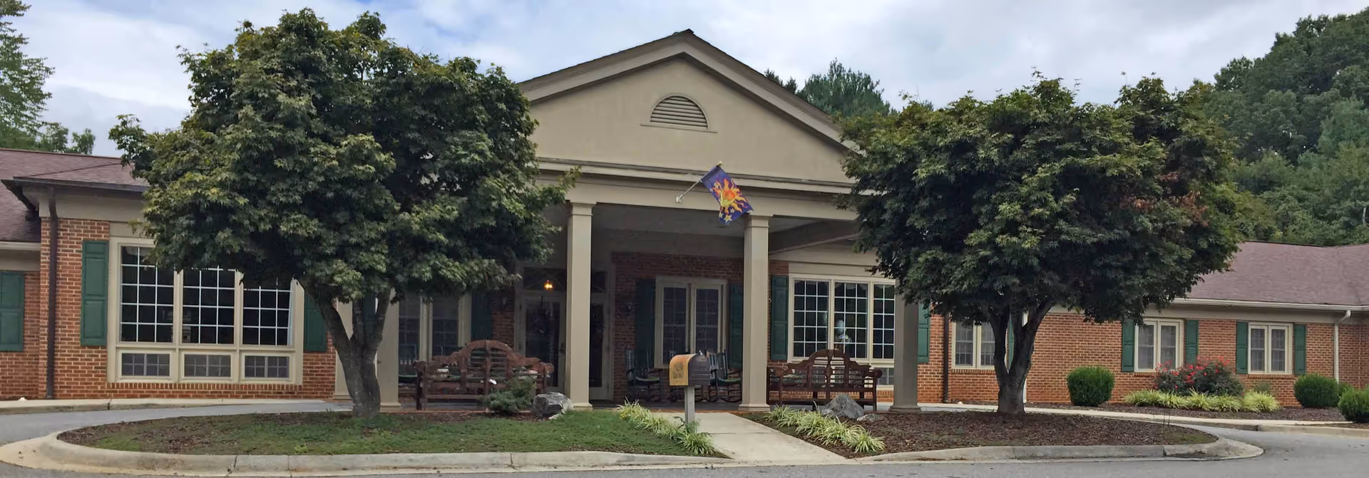 Front entrance of The Elms of Lynchburg senior living building with a covered portico, benches, flag, and trees.