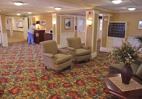 Senior living lounge with patterned carpet, chairs, and a reception desk in the background.