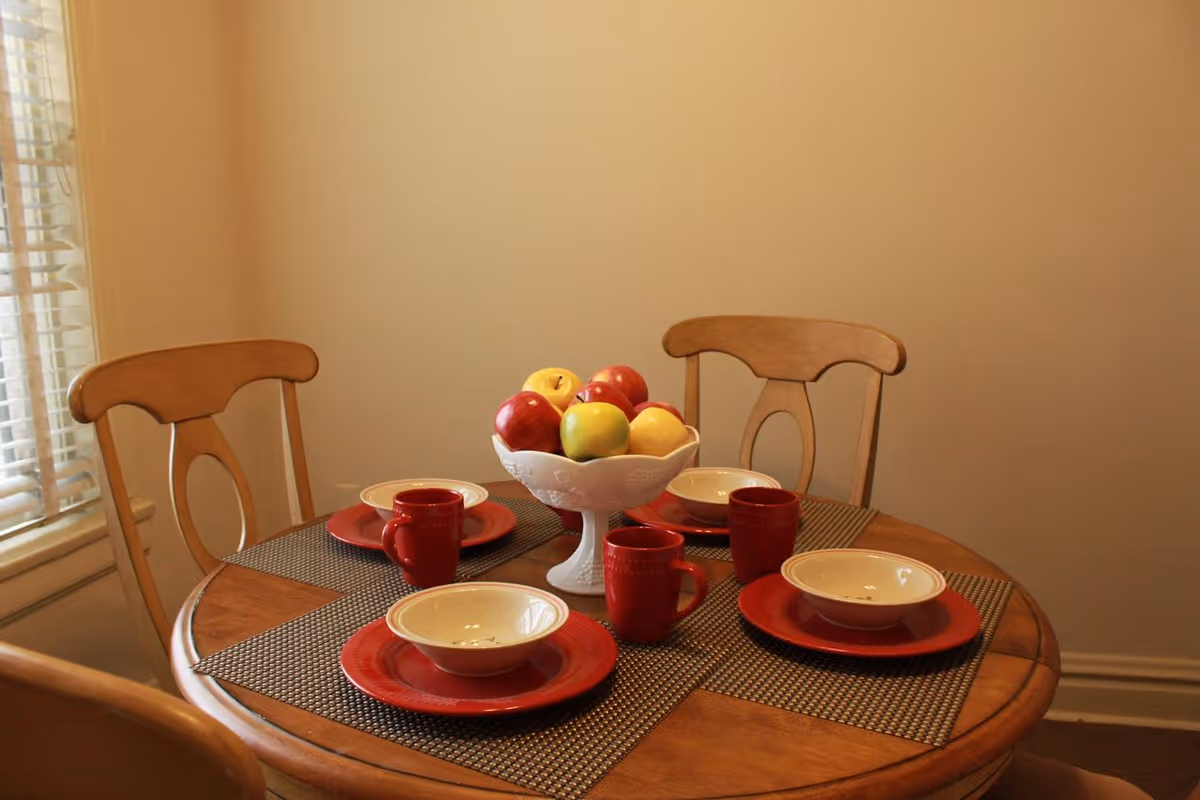 A round wooden dining table set for four with red plates, white bowls, and red mugs. A white pedestal bowl filled with apples and lemons is in the center. Four wooden chairs surround the table, and a window with blinds is visible on the left side.