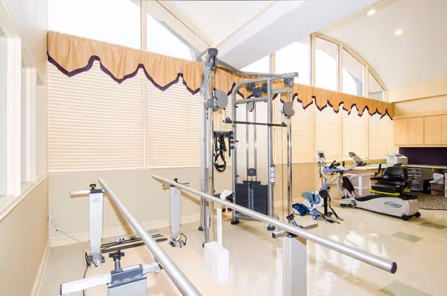 A bright exercise room with large windows covered by beige blinds and valances. The room contains parallel bars for walking support, a multi-functional exercise machine, a recumbent exercise bike, and other fitness equipment. The floor is shiny and light-colored, and the ceiling has recessed lighting.