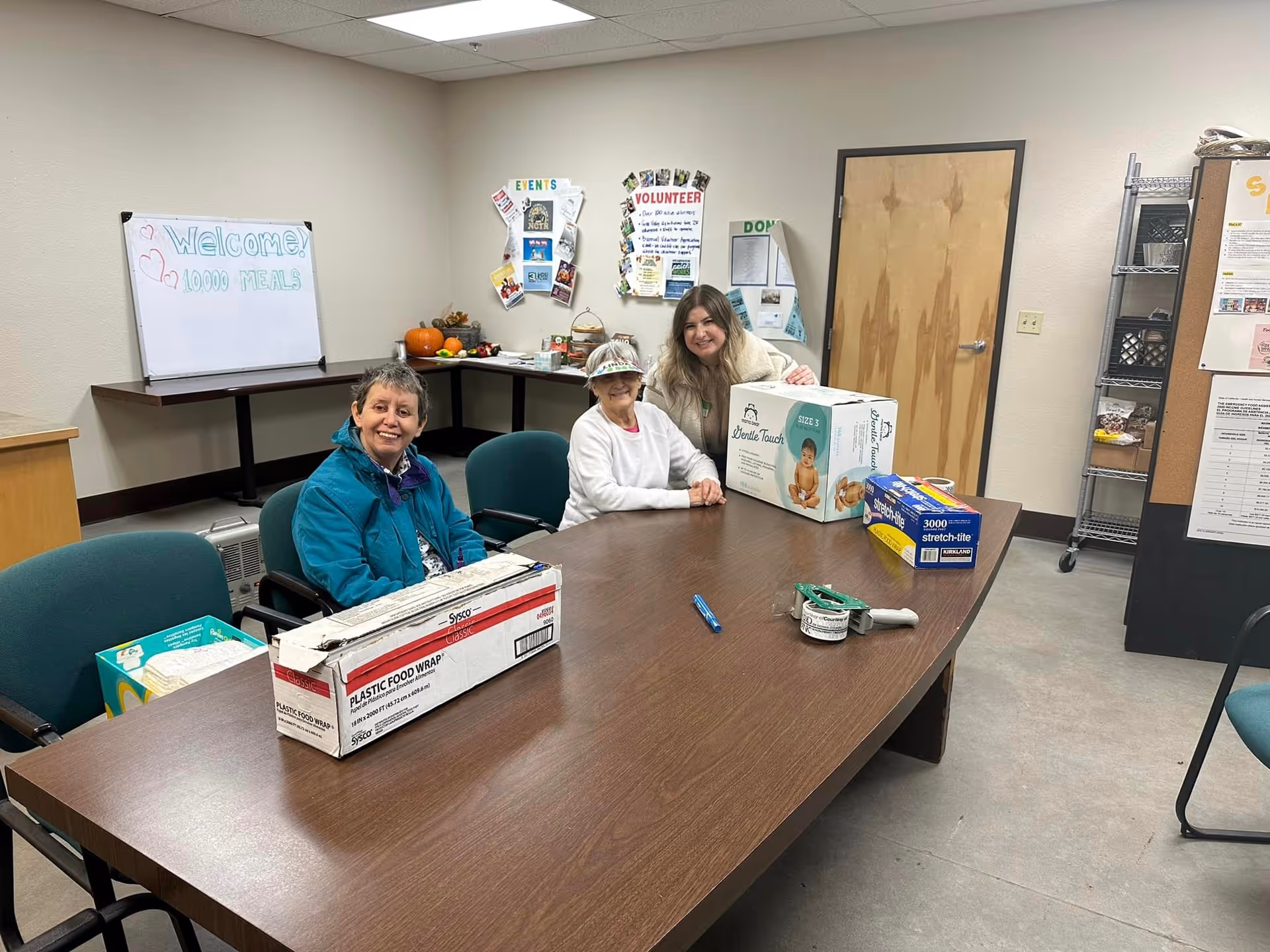Three women sitting around a rectangular wooden table in a meeting room. On the table are boxes of plastic food wrap, disposable gloves, and other supplies. Behind them is a whiteboard with the message 'Welcome! 10,000 MEALS' written on it, and a bulletin board with various notices and decorations. The room has beige walls, a closed wooden door, and metal shelving with additional items.