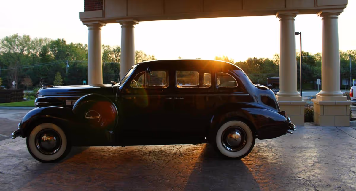A vintage black car with whitewall tires parked under a covered entrance with large columns during sunset, with trees and a parking lot visible in the background.