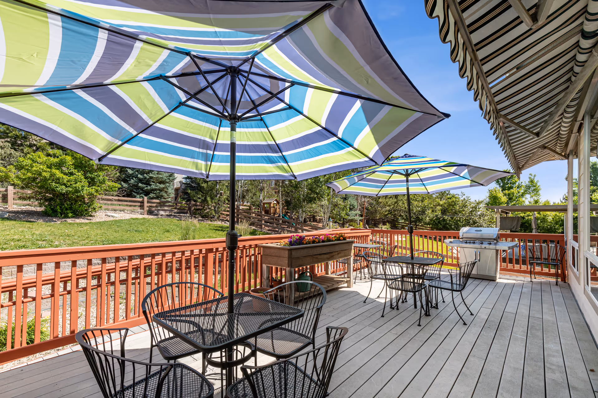 Outdoor deck area with metal tables and chairs, each table shaded by large striped umbrellas. The deck has a wooden railing and a planter box with flowers. There is a grill on the deck and a striped awning attached to the building. Green trees and a clear blue sky are visible in the background.