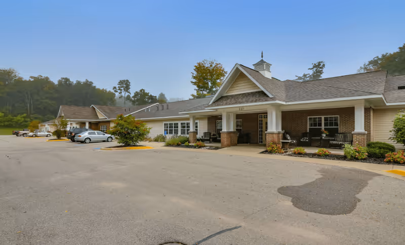 Exterior view of a single-story assisted living facility building with a covered entrance, several parked cars, and surrounding greenery under a clear blue sky.