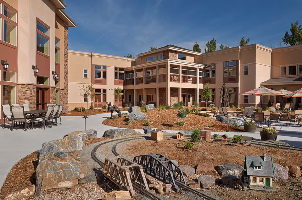 Outdoor courtyard area of a senior living facility with a miniature train track and small model houses surrounded by rocks and mulch. There are patio tables and chairs with umbrellas, and the building has large windows and a beige exterior under a clear blue sky.