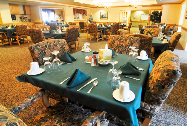 A dining room in a senior living facility with multiple tables covered in green tablecloths, each set with folded napkins, upside-down glasses, cups, and silverware. The chairs have floral patterned upholstery. The room is warmly lit with chandeliers and has a carpeted floor.