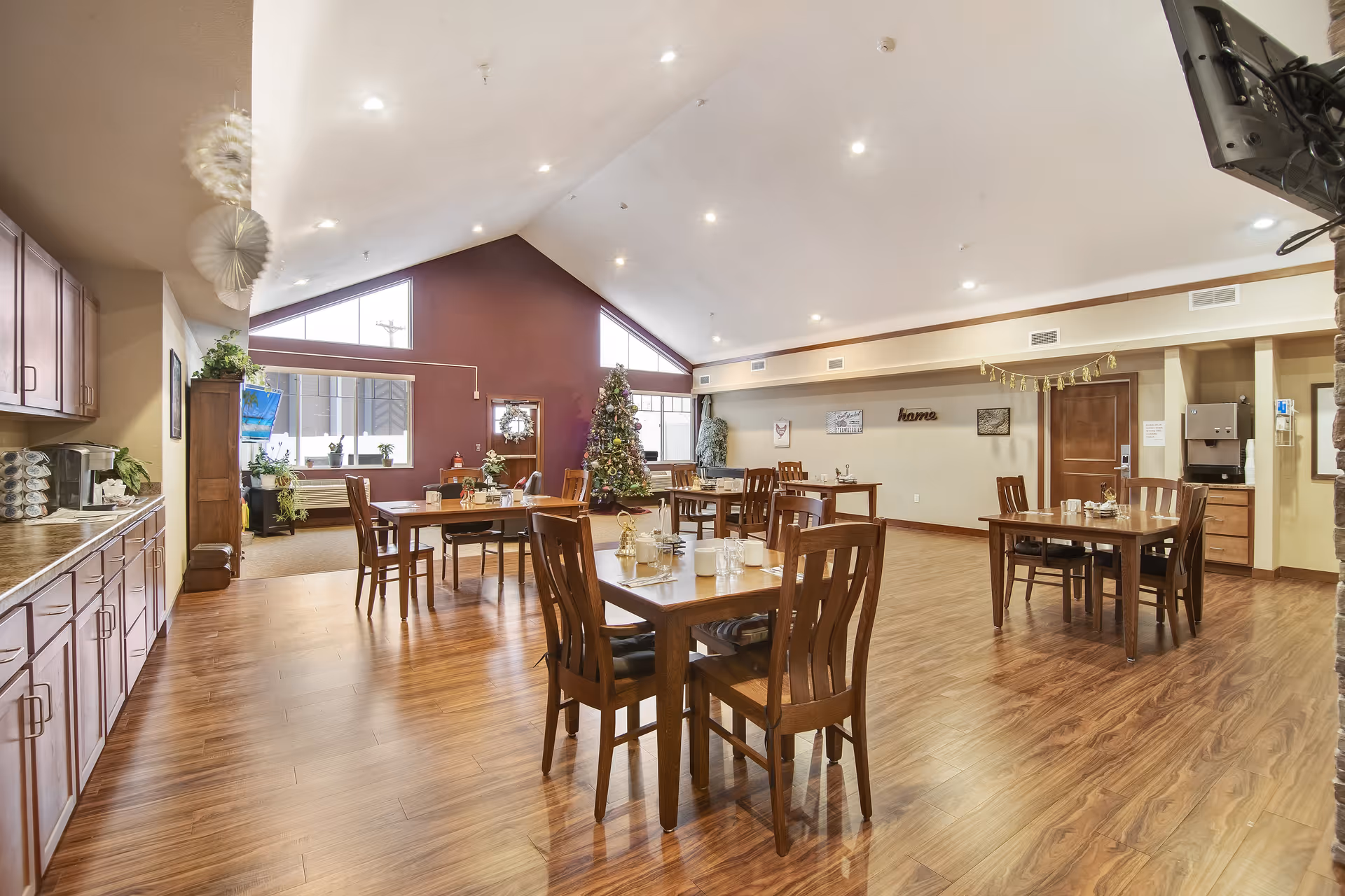 A spacious dining room with wooden floors and several wooden tables and chairs arranged neatly. The room has a high vaulted ceiling with recessed lighting. There is a decorated Christmas tree near the windows, and various plants and decorations are placed around the room. Cabinets and a coffee station are visible on the left side, and a wall with framed pictures and the word 'home' is on the right.
