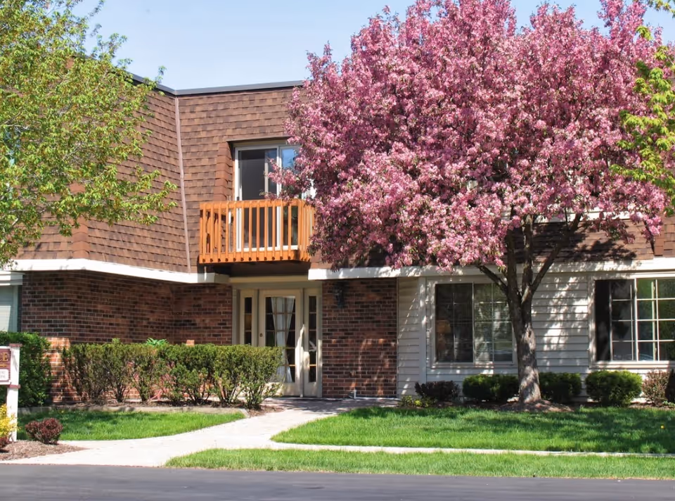 Exterior view of a senior living building with brown siding, a wooden balcony, and flowering tree.