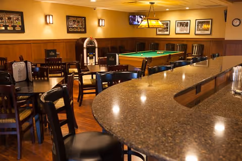 Interior view of a recreational room with a curved granite countertop bar, several wooden chairs and tables, a pool table with balls set up, a jukebox, framed pictures on the walls, and a wall-mounted TV.