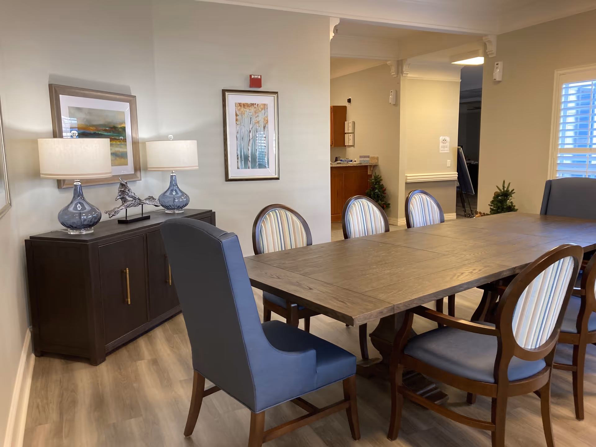 A long wooden dining table with upholstered chairs in a well-lit senior living dining room with a sideboard, lamps, and framed artwork.