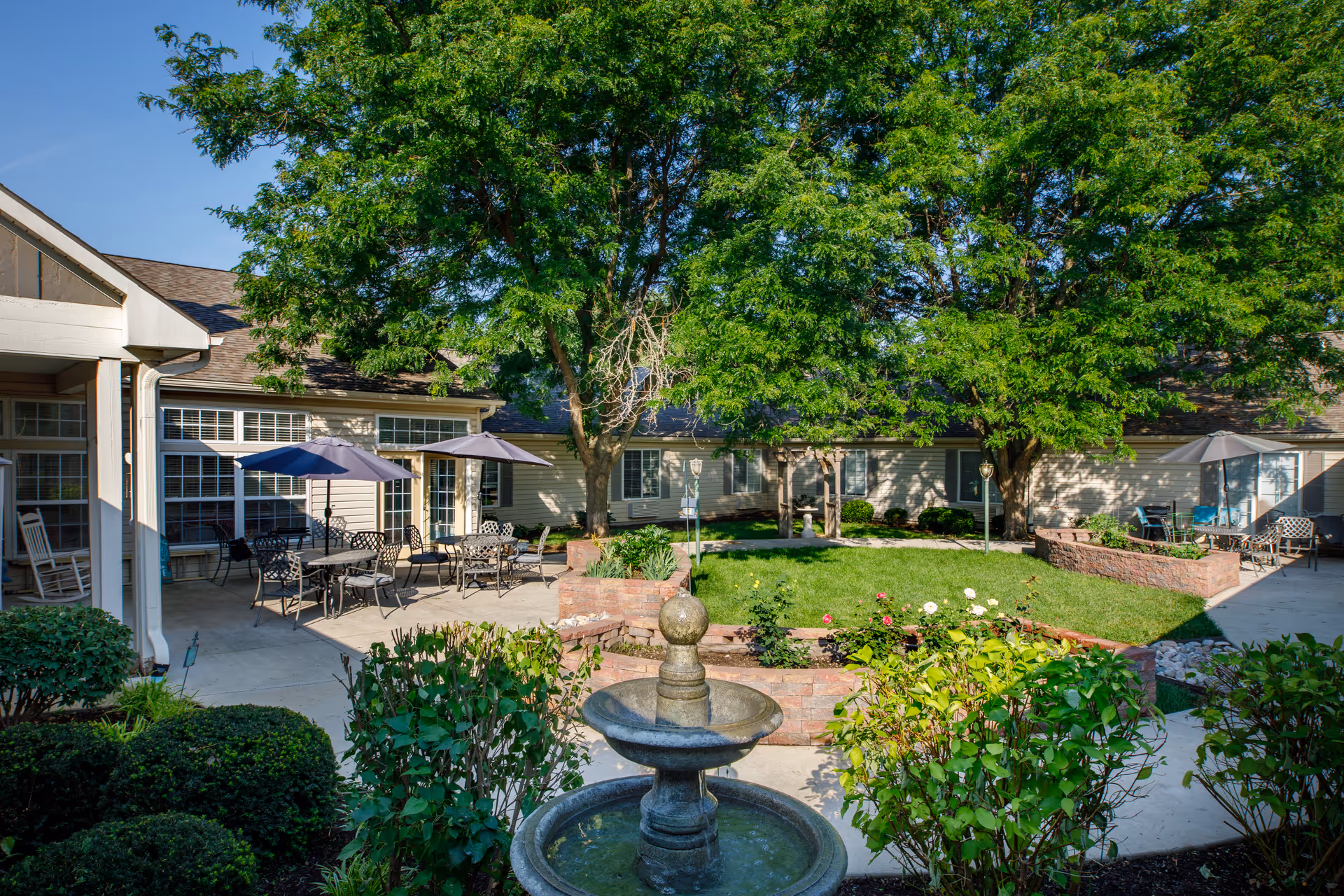 Outdoor courtyard area of an assisted living facility with a central stone water fountain, green bushes, and a grassy area surrounded by brick planters. There are several tables with umbrellas and chairs on the concrete patio, large trees providing shade, and the building with multiple windows in the background.