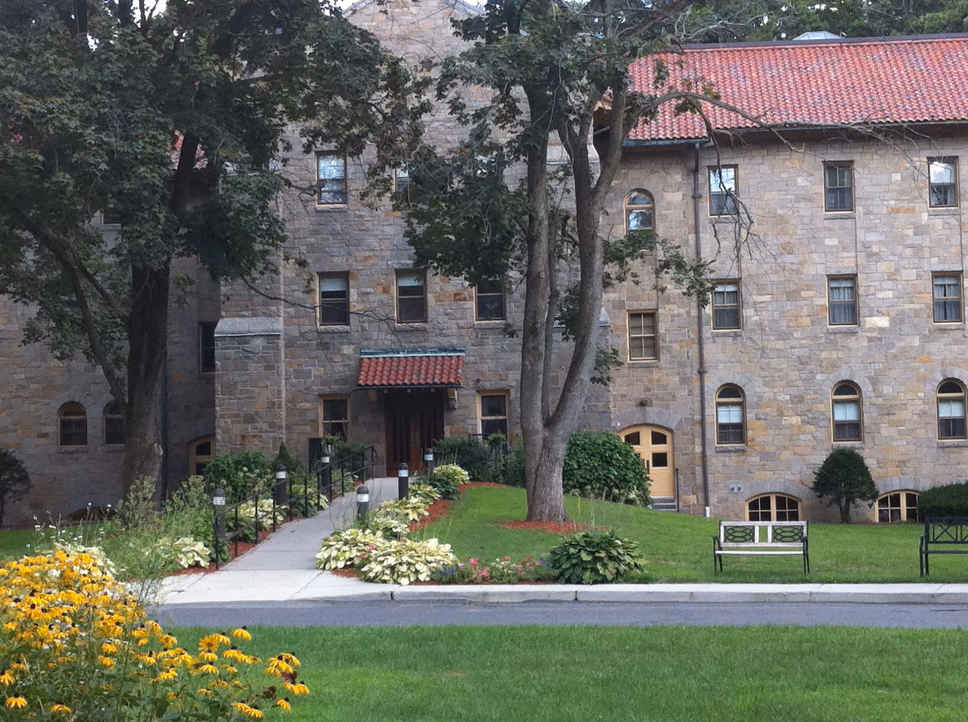 Stone building with multiple windows and a red tiled roof, surrounded by green trees, bushes, and a well-maintained lawn with flower beds and benches. A paved walkway with handrails leads to the entrance of the building.